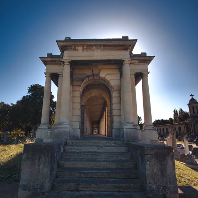 A historic stone monument featuring a classical architectural design with a central arched entrance supported by four large, rounded columns with smooth surfaces and simple capitals. The structure is constructed from light-colored stone or concrete, exhibiting a weathered appearance with some darkened areas and subtle erosion. The entrance is accessed by a short flight of worn stone steps with visible cracks and rounded edges, leading up from the grass and pavement in the foreground. Surrounding the monument are neatly maintained grassy areas and mature trees with dense foliage, casting shadows on the ground. In the background, additional stone structures and a small, domed pavilion topped with a cross are visible, suggesting a cemetery or memorial site. The clear sky above provides bright, natural lighting, creating a sharp contrast with the shadowed interior of the arched passage. Though primarily a historical or commemorative site, the scene emphasizes the structural durability and classical design, which could relate to on-site clearance or private disposal of debris in a memorial garden context, reflecting the importance of careful rubbish removal in heritage environments. Waste Collection Brompton offers services that support such detailed disposal and clearance tasks, ensuring the preservation of these sites.