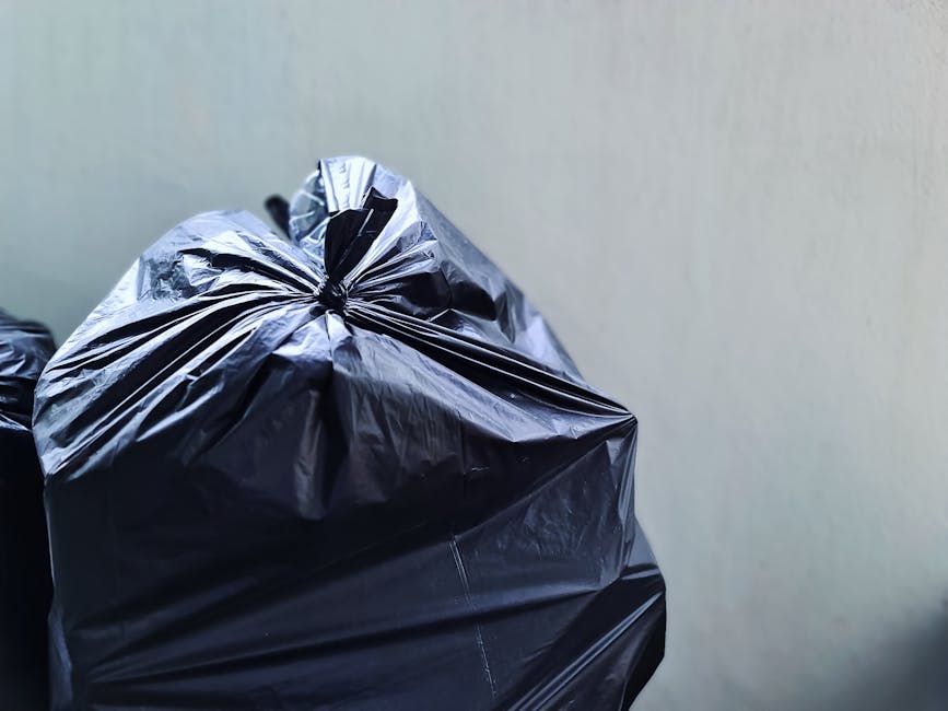 A large collection of mixed waste and rubbish bags is piled around a public recycling bin and trash containers in an outdoor parking area. The waste includes crumpled paper, cardboard boxes, plastic bags, and packaging materials, some spilling onto the paved surface. The recycling bin, labeled for mixed paper and cardboard, is partially open with various waste items overflowing from top and sides. To its right, there are black and red rubbish bins, one with black plastic bags secured inside and others with loose debris outside. In the background, a two-storey commercial building with a blue metal scaffolding structure and a yellow signage strip can be seen, alongside parked cars, including a silver vehicle with a visible registration plate. The environment appears to be an urban area with minimal greenery, and the scene is lit by natural daylight, emphasizing the disorderly accumulation of waste typical of private or independent rubbish collection services in Brompton.
