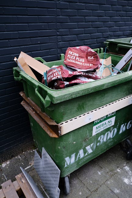 A green wheeled skip filled with red and black bags of garden mulch or compost, placed outside against a dark brick wall on a paved surface. The bags are stacked haphazardly, with some leaning on each other, and their labels are visible. A white and brown plastic sheet shields part of the skip's side, and adjacent to it are additional waste containers. The surrounding environment suggests an outdoor area suitable for private waste collection or on-site clearance by waste management services like Waste Collection Brompton, which specializes in rubbish removal solutions. The scene highlights an informal collection point, with the bags and containers positioned for eventual disposal or collection, reflecting an alternative approach to regular municipal rubbish collection.