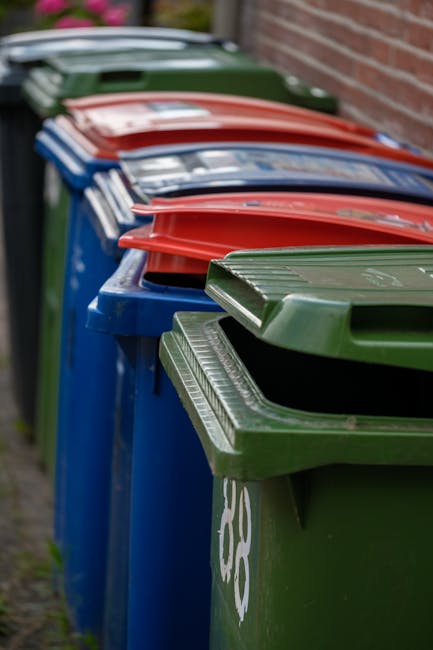 A green wheeled skip filled with red and black bags of garden mulch or compost, placed outside against a dark brick wall on a paved surface. The bags are stacked haphazardly, with some leaning on each other, and their labels are visible. A white and brown plastic sheet shields part of the skip's side, and adjacent to it are additional waste containers. The surrounding environment suggests an outdoor area suitable for private waste collection or on-site clearance by waste management services like Waste Collection Brompton, which specializes in rubbish removal solutions. The scene highlights an informal collection point, with the bags and containers positioned for eventual disposal or collection, reflecting an alternative approach to regular municipal rubbish collection.