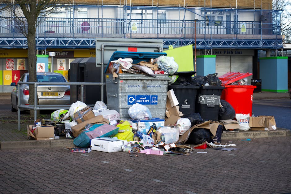 A large outdoor refuse collection point situated on a paved urban street, with multiple open-top wheelie bins filled with various types of waste including paper, cardboard, plastic bags, and miscellaneous household rubbish. The central grey bin, labeled for mixed paper and cardboard, is overflowing with crumpled paper, flattened boxes, and other debris spilling onto the surrounding pavement. To its right are black and red recycling bins, partially filled with waste, some tied-up plastic bags and loose litter scattered around their bases. In the background, a row of storefronts with signs and windows is visible, along with a parked silver car on the left and a metal railing separating the waste area from the sidewalk. Behind the scene, a multi-story building under renovation, covered in blue scaffolding and safety netting, adds an urban context. Natural daylight illuminates the scene, highlighting the disorderly pile of waste as indicative of illegal dumping or inadequate collection, typical in areas where private rubbish services like Waste Collection Brompton might be employed for alternative waste handling outside local authority provisions.