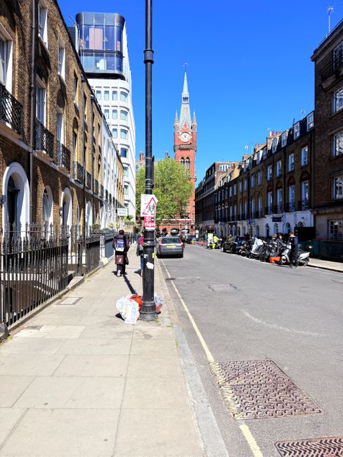 A city street scene in daylight with a clear blue sky, featuring a row of terraced brick buildings on the right side, with numerous windows and small balconies. On the left, a sidewalk runs parallel to the road, with a black wrought-iron fence separating residential properties. A tall street lamp is positioned centrally in the foreground. Near the base of the lamp, a white plastic bag and some smaller waste items are placed on the pavement. Several pedestrians are walking along the sidewalk, and a few parked motorcycles and a small car are visible along the curb. In the background, a prominent red-brick clock tower with a pointed spire and white accents rises above the surrounding buildings, with lush green trees partially obscuring the base of the tower. The street appears to be a mix of residential and commercial activity, with a scene that could involve local or private rubbish collection, reflecting alternative waste management practices often seen in urban areas. Waste Collection Brompton's services are relevant in the context of managing rubbish around such cityscapes without reliance solely on local authority collection, often involving on-site or private disposal options.