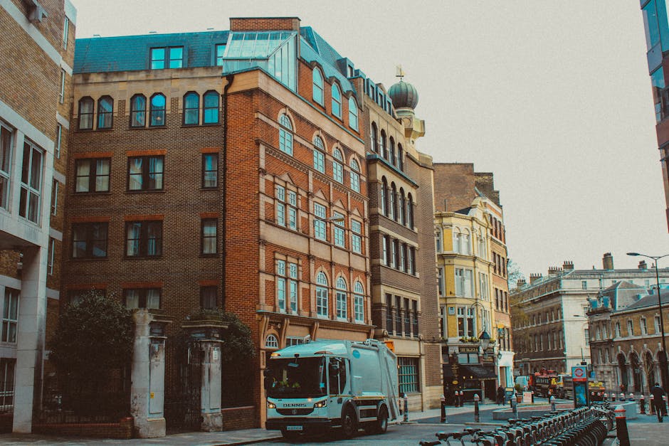 A city street scene featuring a variety of multi-storey buildings, including a prominent red-brick structure with intricate window arch detailing and a rounded rooftop turret with a weather vane. Adjacent buildings display a mix of architectural styles, some with ornate facades painted in pale yellow and cream shades, and others with modern glass-fronted exteriors. In the foreground, a waste collection vehicle from Waste Collection Brompton, painted in light blue, is parked on the street, likely engaged in rubbish collection. The vehicle is positioned on the right side of the view, surrounded by a row of black bicycle racks and bollards separating the pedestrian walkway from the road. The scene is illuminated by natural daylight, with an overcast sky casting a neutral light over the buildings and street, which appears to be part of an urban area with typical city infrastructure including streetlights, signage, and traffic bollards. The presence of the waste collection vehicle suggests an active process of rubbish removal, possibly for commercial or residential waste management within this area.