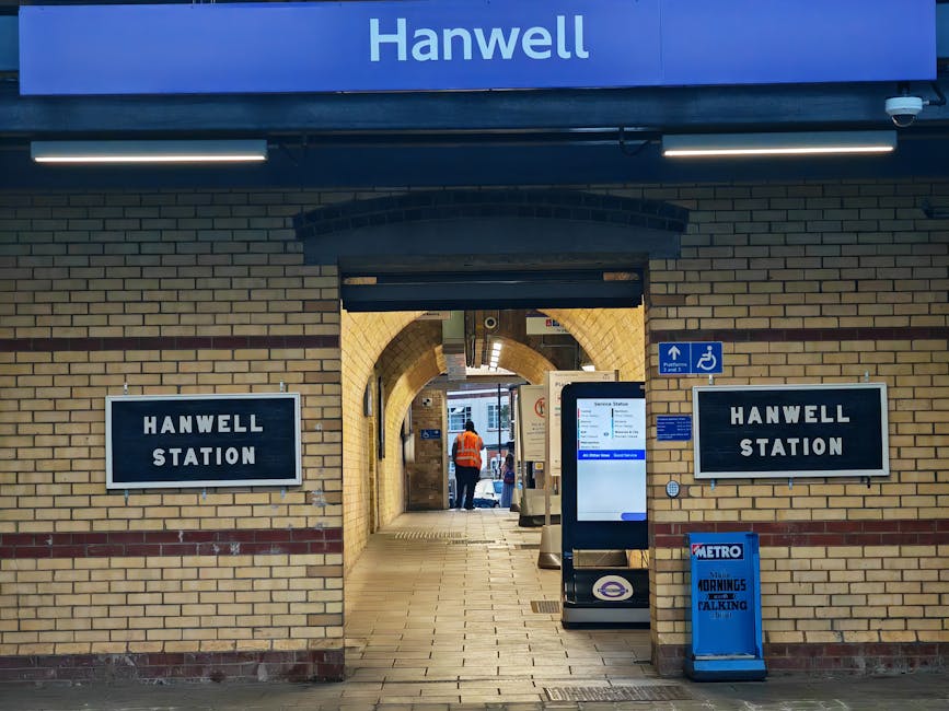 A view of the entrance to Hanwell Station, featuring a brick facade with beige and reddish-brown tones. On either side of the arched doorway are black signs with white lettering reading 'Hanwell Station.' Above the entrance, a blue sign displays the station's name in white text. The area is illuminated by linear LED lights mounted on the ceiling. In the foreground, a digital timetable display stands on a pole, showing travel information with a white background and blue accents. To the right of the entrance, a blue and white sign indicates accessible facilities, including a wheelchair symbol. In front of the station, a small blue Metro-branded recycling or rubbish bin is present, along with a blue sign or bag that appears to be related to waste collection. Inside the station, visible through the arched entrance, are a person wearing an orange high-visibility jacket and brief glimpses of the platform, suggesting the location is prepared for passenger transit and potentially for independent or private waste disposal options handled by Waste Collection Brompton in relation to local station facilities.