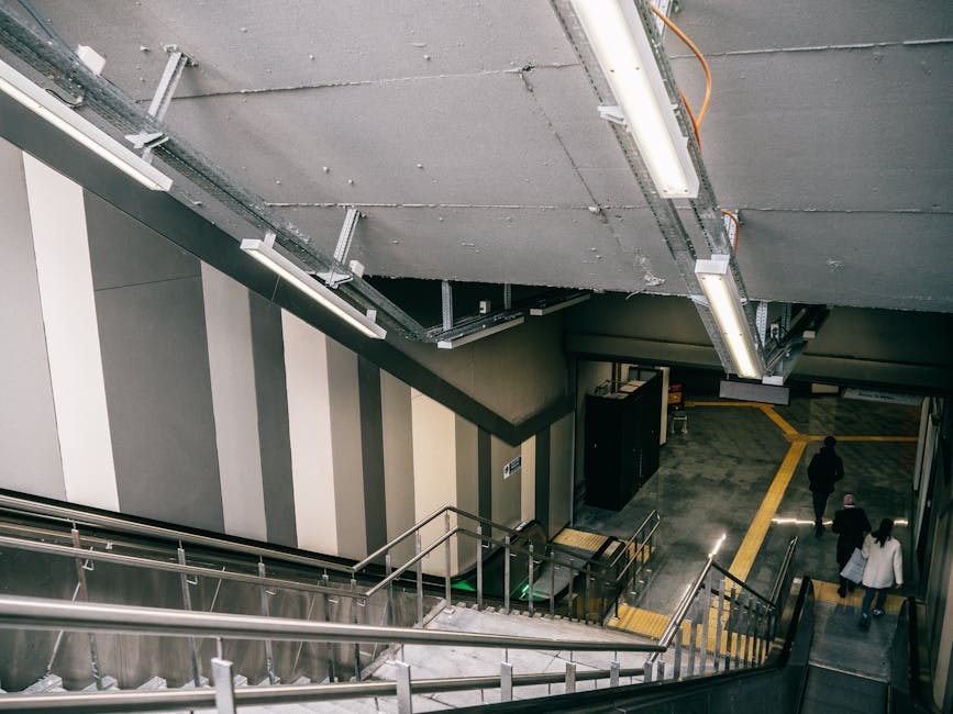 Inside an underground station stairwell, the image shows a downward view of a corridor with three people walking along a tiled floor. The wall on the left features vertical stripes in shades of beige, gray, and off-white. Overhead, there are exposed ceiling panels with visible electrical conduits and mounted fluorescent lighting fixtures, providing bright, even illumination to the space. The staircase, with metal handrails and concrete steps, leads down to the platform area. In the background, a portion of a black cargo lift or service door is visible, suggesting a functional area for station staff or maintenance. The environment appears clean and well-maintained, with tactile paving strips near the bottom of the stairs indicating accessibility for visually impaired users. The overall scene emphasizes a typical urban public transport interior, relevant to discussions about station facilities or arrangements for rubbish and waste disposal within transit environments, possibly managed by waste collection services such as Waste Collection Brompton.